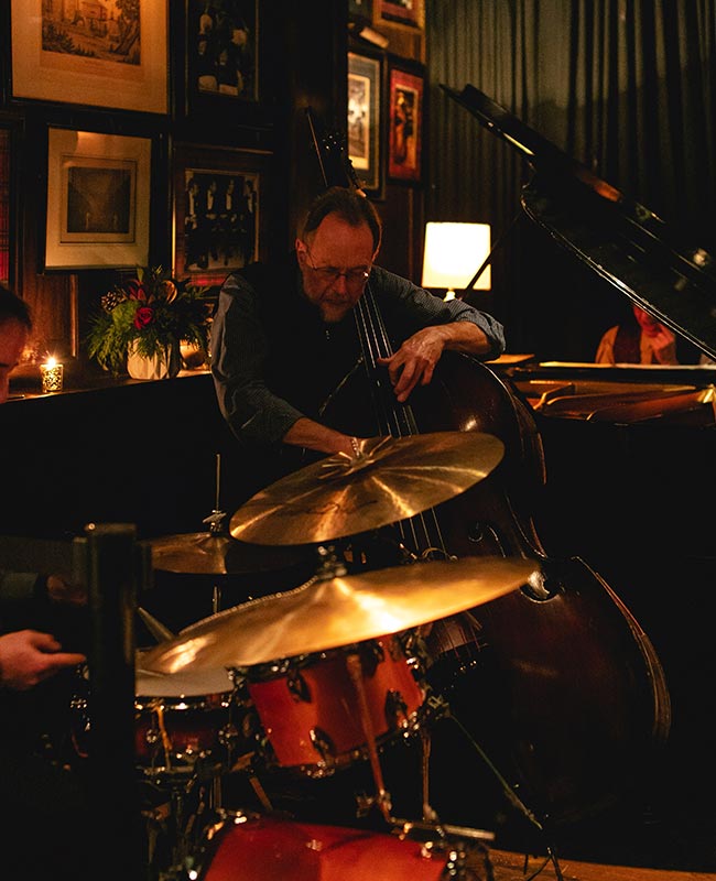 A jazz musician plays the standup bass in a lounge with drums in the foreground and a piano in the background