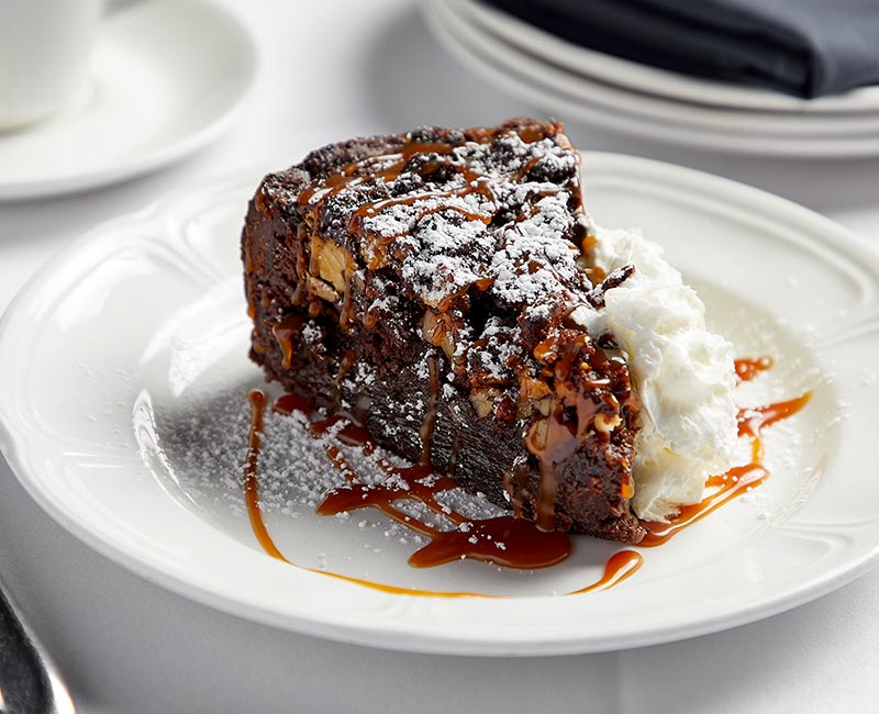 A decadent brownie cake sits on a plate with whipped cream, caramel, and powered sugar as a garnish